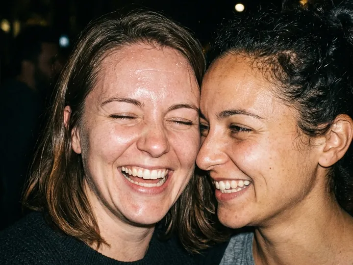 Two friends cheek to cheek, laughing, lit by a camera flash