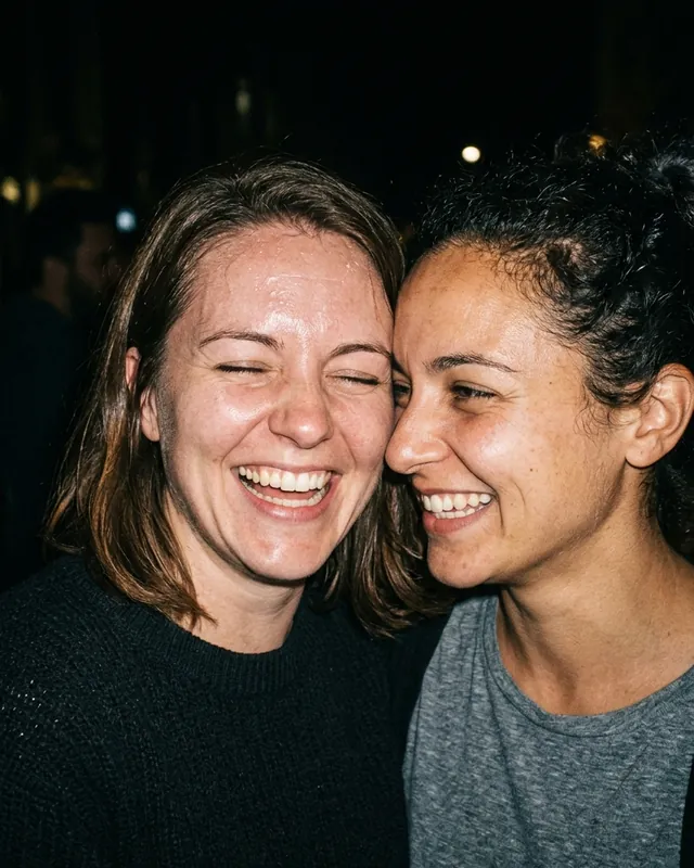 Two friends cheek to cheek, laughing, lit by a camera flash